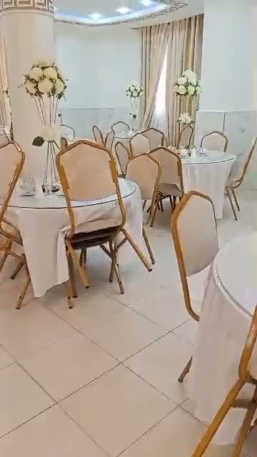 Salle dressée en réception : tables rondes nappées de blanc, chaises dorées et hautes compositions de roses blanches